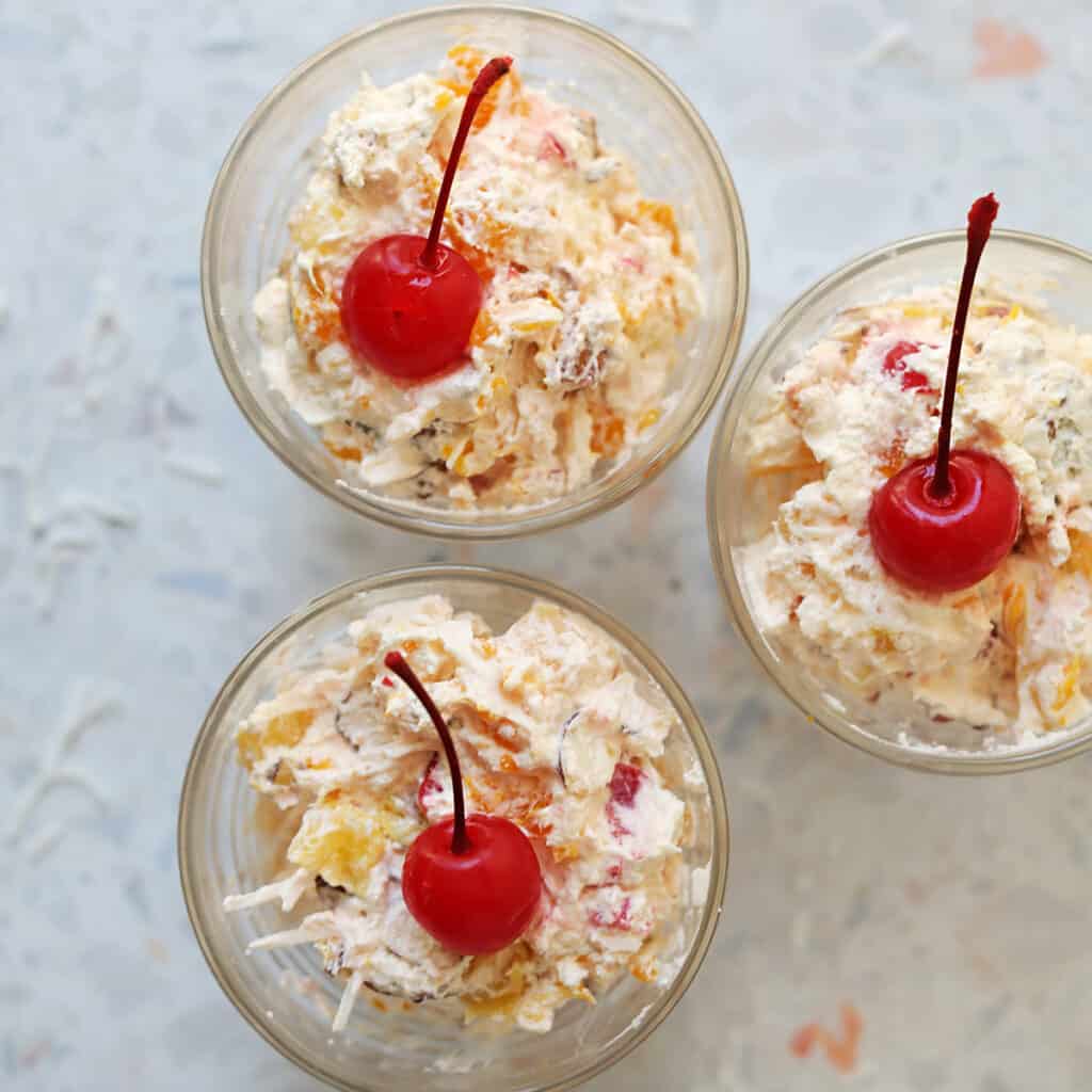 creamy ambrosia fruit salad in three small cups with a cherry on top